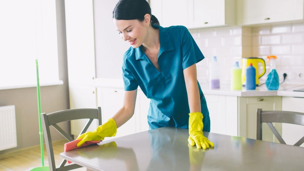 A woman wearing yellow gloves and a blue uniform cleans a kitchen table with a pink cloth. Cleaning supplies are visible in the background, showcasing professional cleaners ready for any move out cleaning in San Mateo.