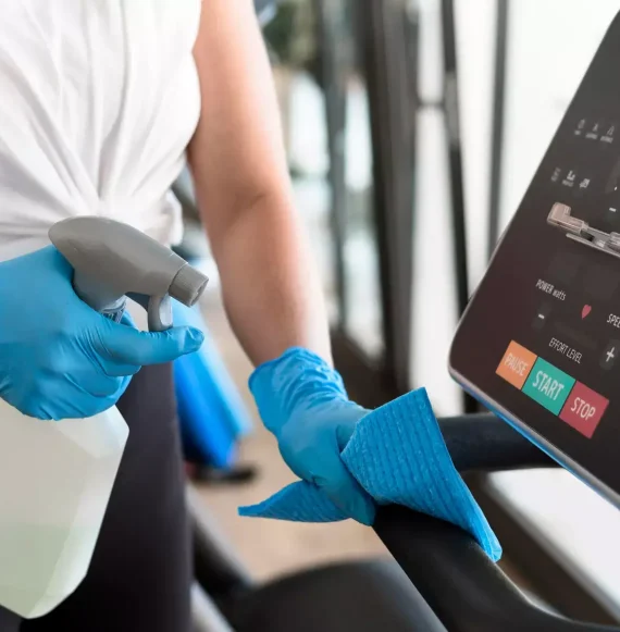 A person wearing blue gloves cleans a gym treadmill with a spray bottle and cloth, focusing on the console and handrail—demonstrating the thoroughness of San Mateo cleaners.